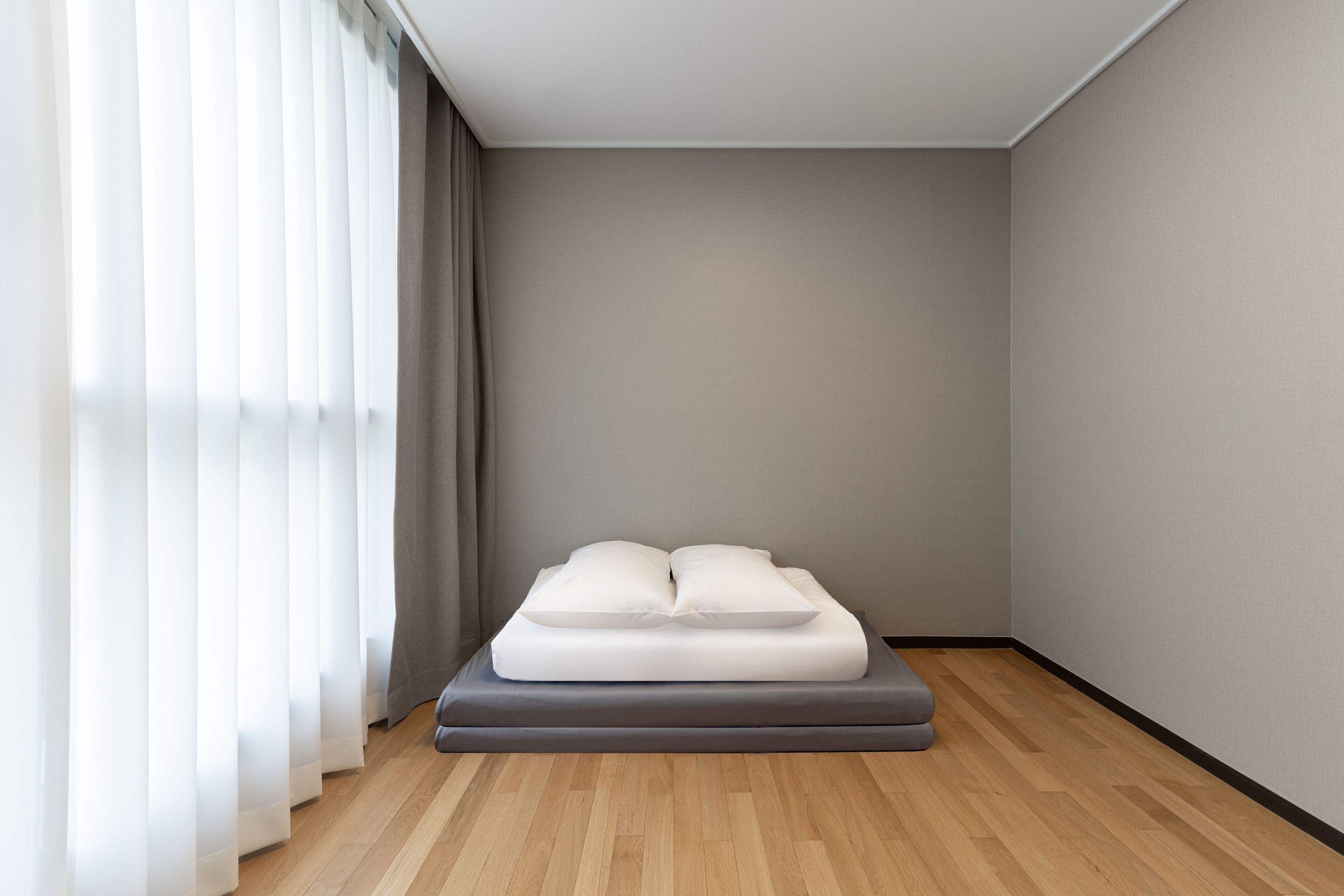 A minimalist bedroom with a single bed, light wooden flooring, and sheer curtains.