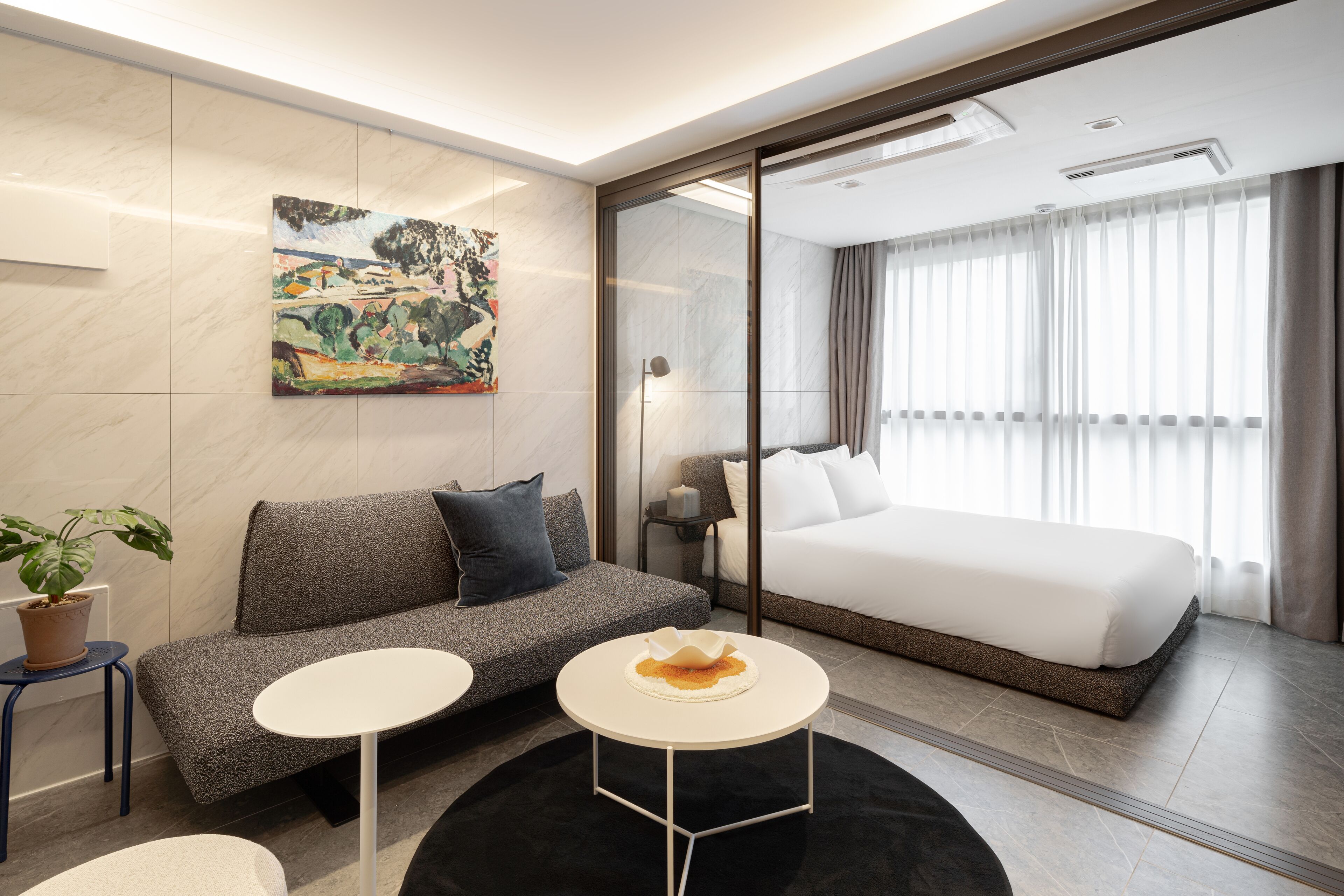 A modern hotel room with a grey sofa, a white bed, a small round table, and a painting on the wall.