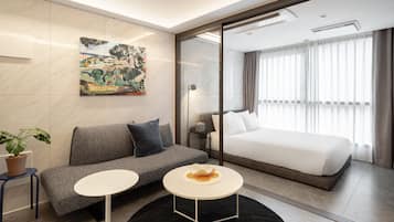 A modern hotel room with a grey sofa, a white bed, a small round table, and a painting on the wall.