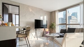 A modern hotel room with a flat-screen TV, a dining area with a chair and table, a sofa, a potted plant, and a view of the cityscape through large windows. - Le Collective Gangnam SAC ROIUM (Seoul)