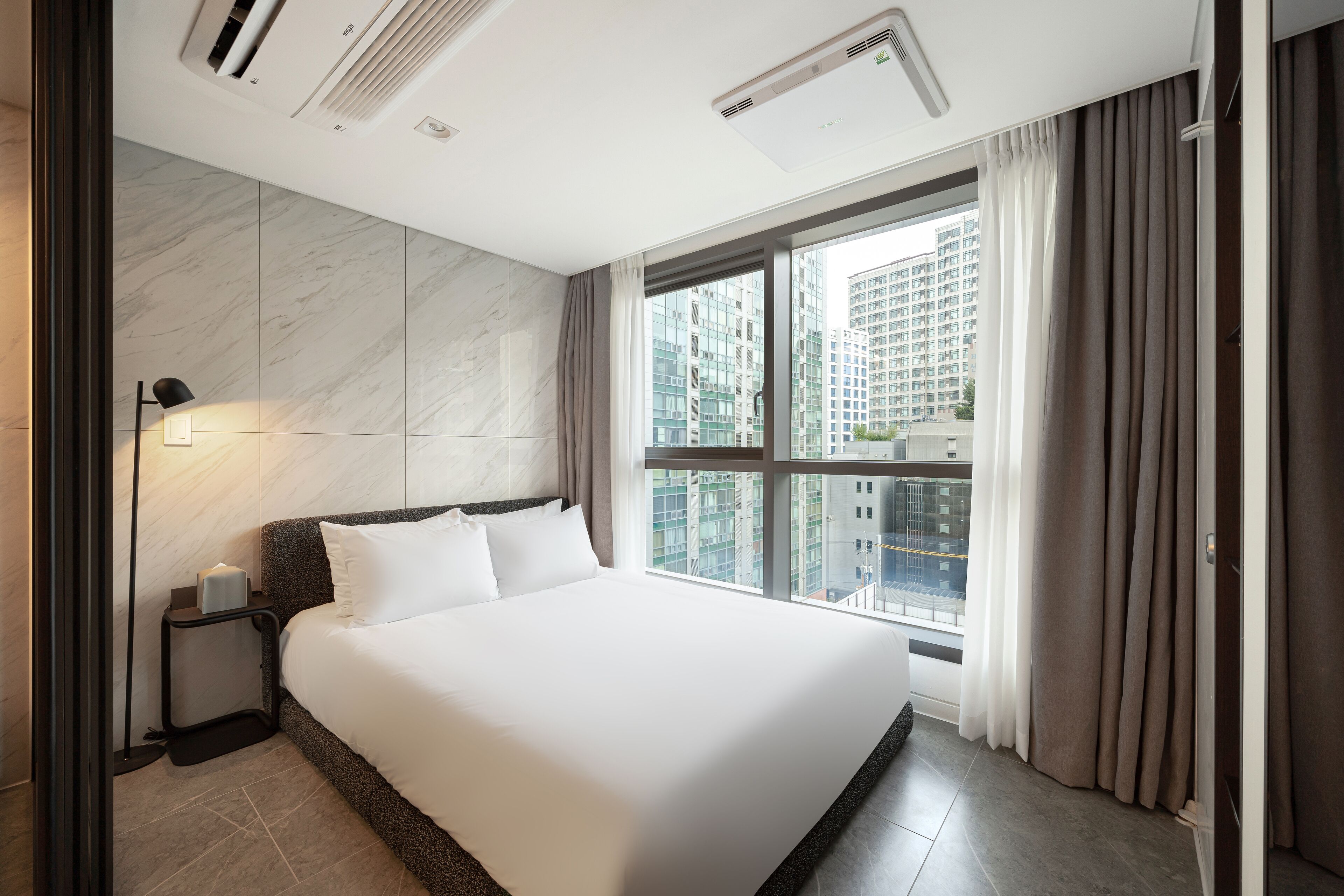 A modern hotel room with a large bed, a nightstand, a lamp, and a view of tall buildings through a large window.