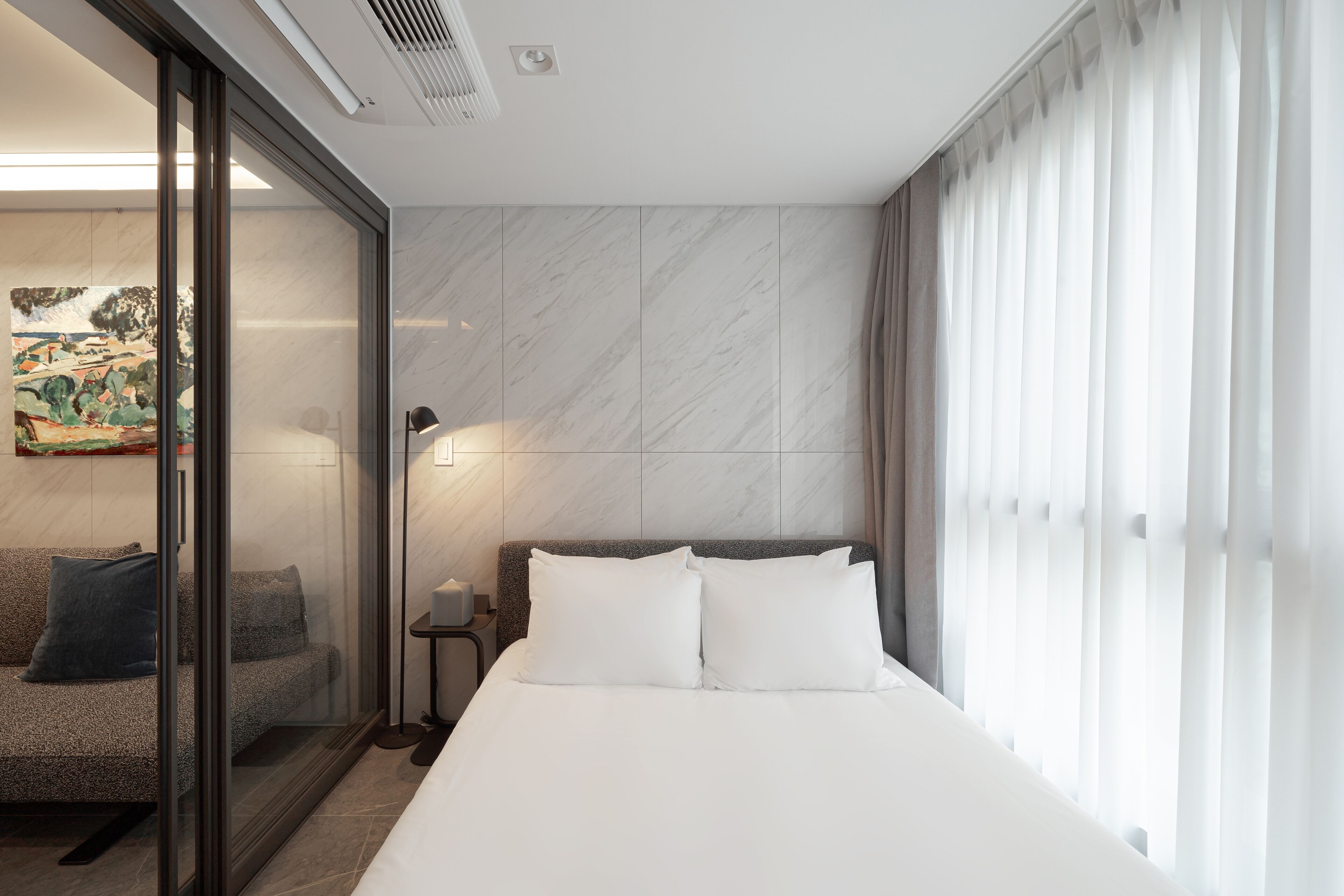 A hotel room with a bed, a nightstand, a lamp, a painting on the wall, and a sliding glass door leading to a seating area.