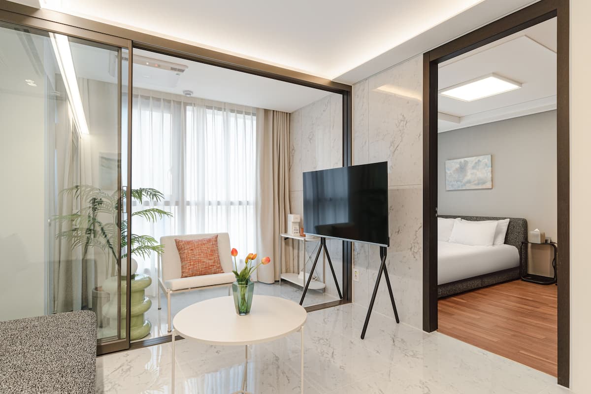 A modern hotel room with a glass partition, a sofa, a small table with a vase, a flat-screen TV on a stand, and a bed with a grey headboard.
