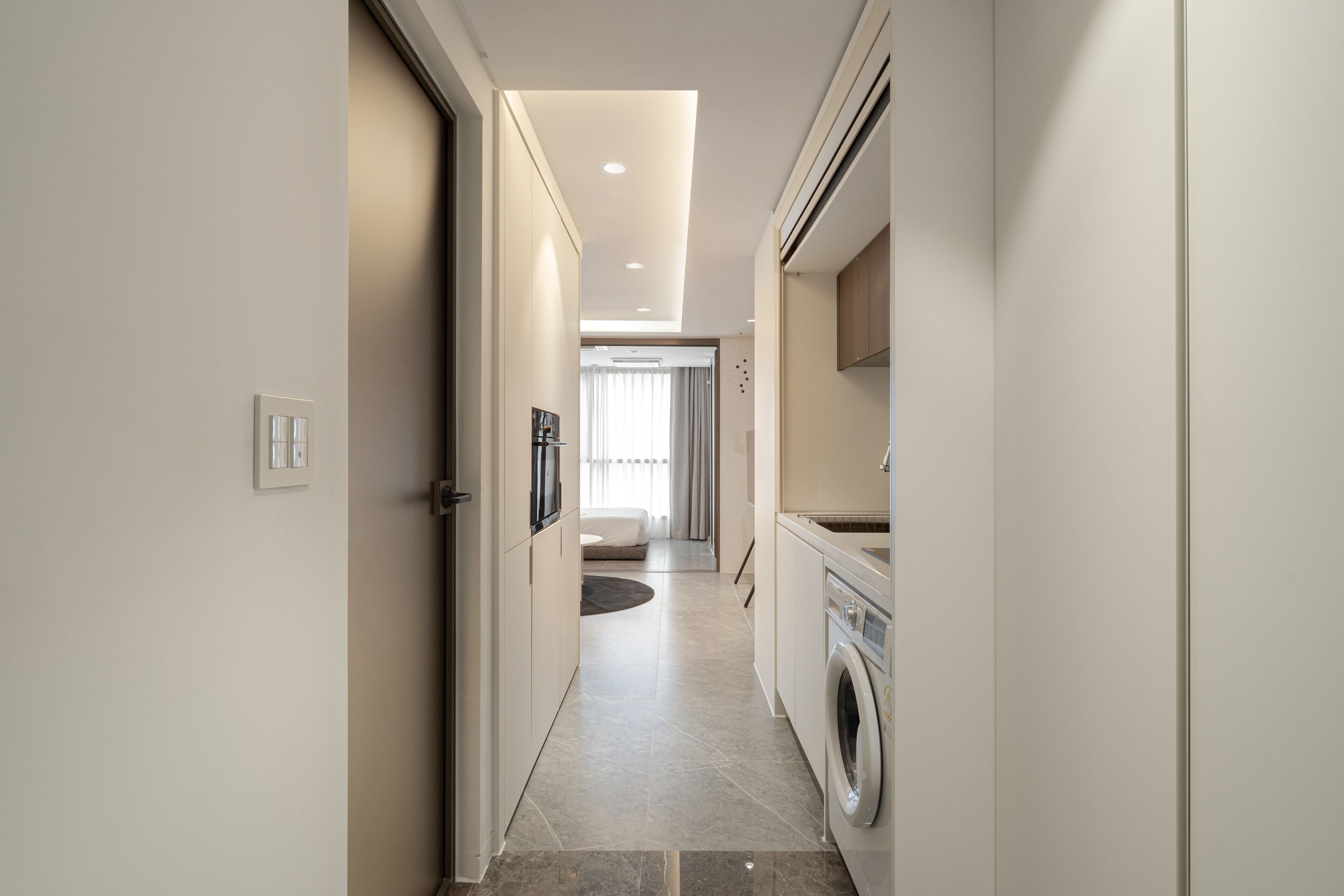 A modern, narrow hallway with a washing machine, a kitchen area, and a living space visible in the distance.