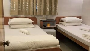 Une chambre d’hôtel avec deux lits simples, une petite table, une table de chevet, un miroir, une fenêtre avec des rideaux et un ventilateur de plafond.