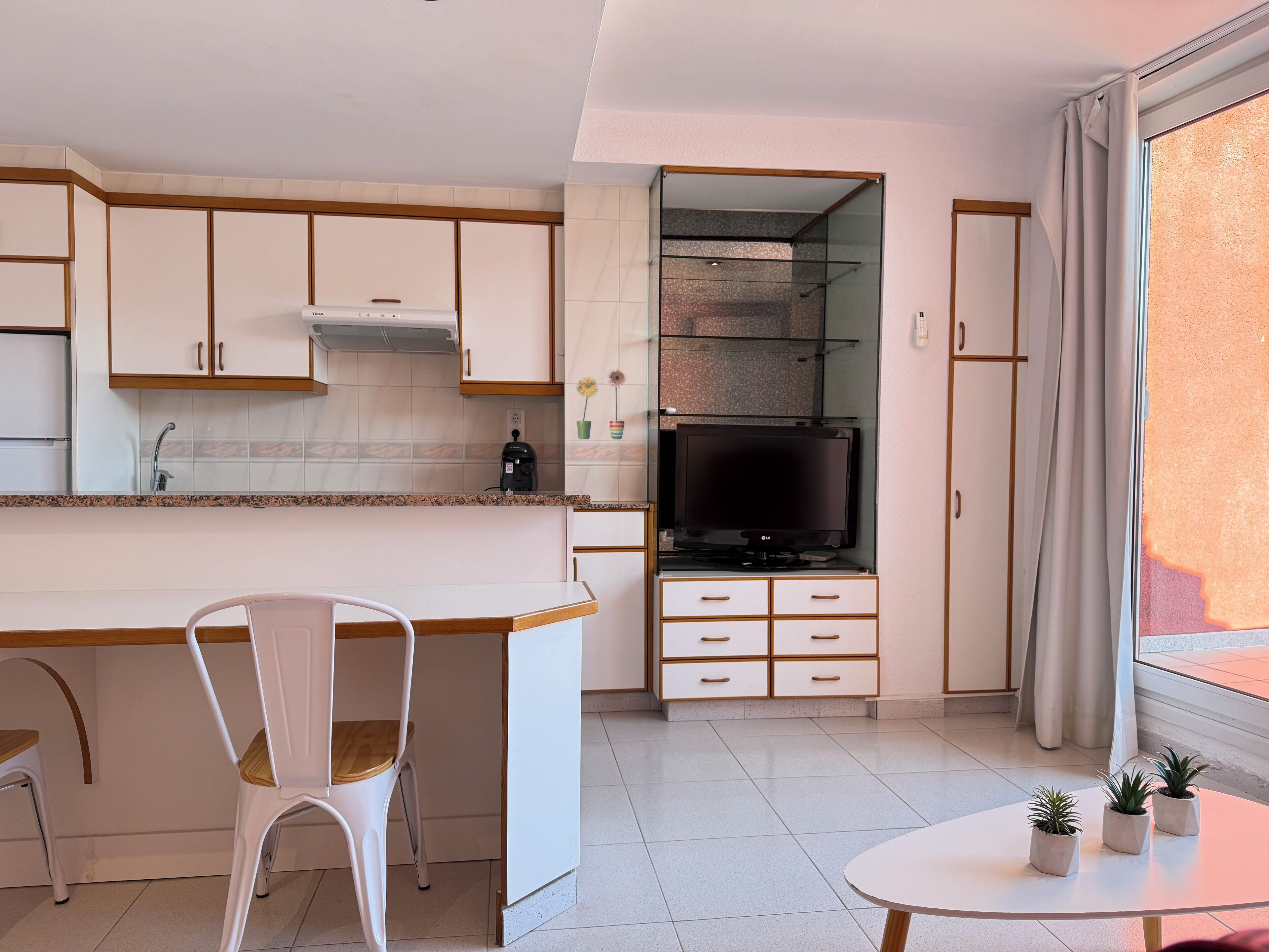 Una cocina moderna con armarios blancos, estufa integrada y un televisor sobre un mueble bajo. Hay un comedor con una silla blanca y una pequeña mesa con plantas en macetas.