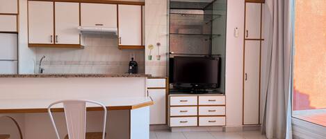 Una cocina moderna con armarios blancos, estufa integrada y un televisor sobre un mueble bajo. Hay un comedor con una silla blanca y una pequeña mesa con plantas en macetas.