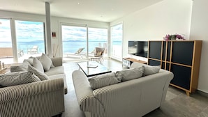 A modern living room with striped sofas, a glass coffee table, and a TV on a wooden stand. Large windows offer a view of the sea. - Agi Turo Villa 1 Casa for rent in Roses Girona. (Roses)