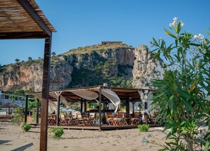Un ristorante sulla spiaggia con posti a sedere all'aperto, vista su una scogliera e un edificio in cima.