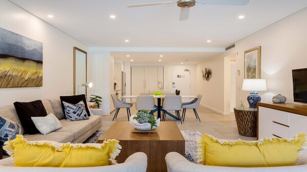 A modern living room with a sofa, coffee table, and yellow cushions. There is a dining area with a table and chairs, and a kitchen in the background. - Azure 187 | Port Douglas (PORT DOUGLAS)