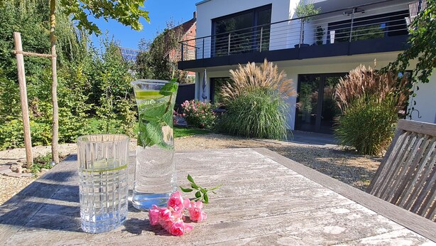 A wooden table with two clear glasses, a small vase with a plant, and pink flowers. - Place2be - Your feel-good apartment, Ankommen und wohlfühlen! (Calden)
