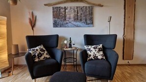 A room with two black armchairs, a small round table with a bottle and glass, a painting of a snowy forest, a hanging plant, and a wooden beam on the wall. - Place2be - Your feel-good apartment, Ankommen und wohlfühlen! (Calden)