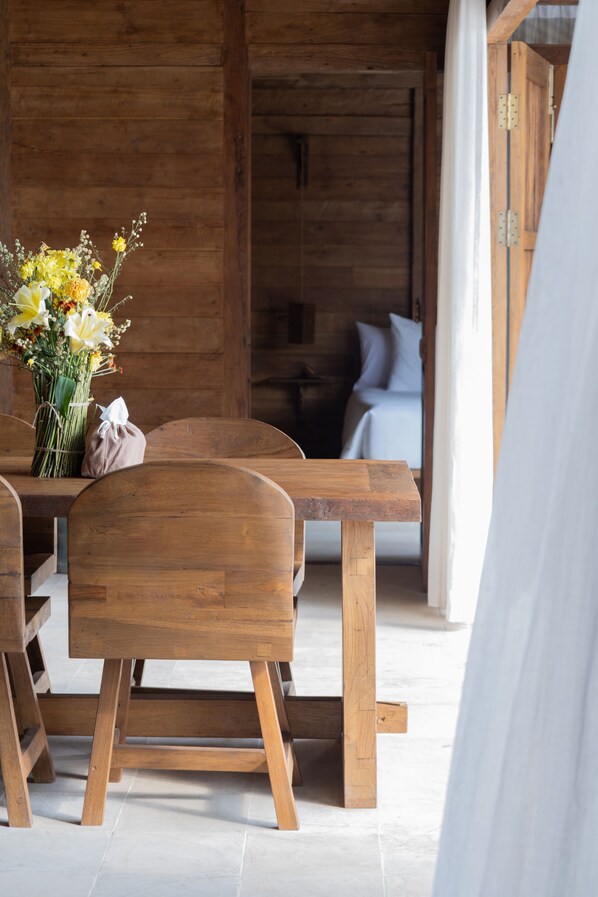 Une table à manger en bois, entourée de chaises, un vase de fleurs et un lit à l’arrière-plan.