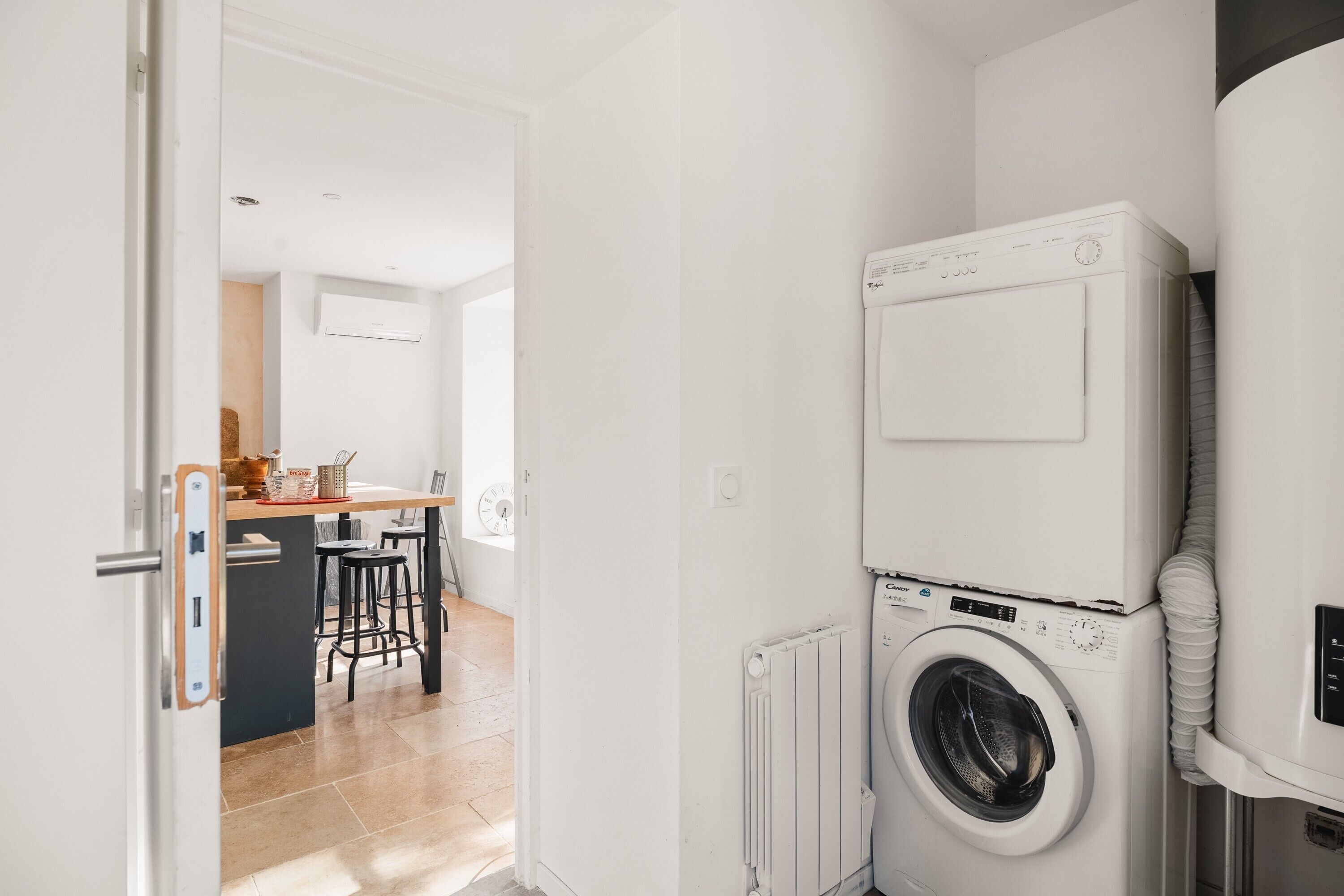 Un espace buanderie avec une machine à laver et un sèche-linge superposés, un coin cuisine avec une table et des tabourets, et une porte visible donnant sur une autre pièce.