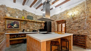 Une cuisine rustique avec un mur en pierre, des poutres en bois, un grand îlot central avec des tabourets et un porte-ustensiles suspendu.