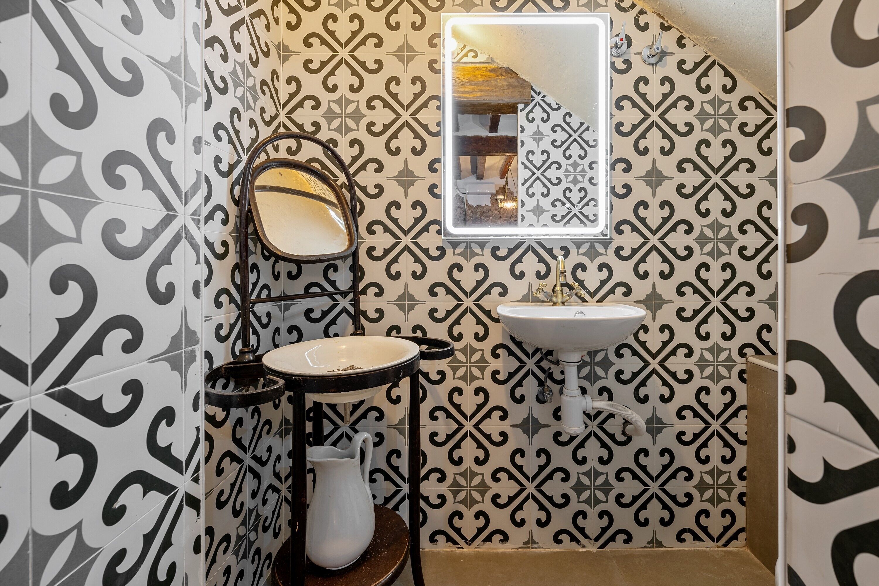 Une salle de bain avec des carreaux décorés, un miroir, un lavabo et un pichet posé sur un tabouret.