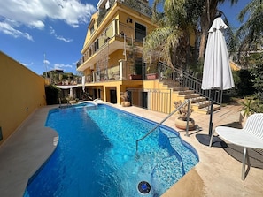 A swimming pool with a clear blue water, a white umbrella, and a multi-story building with balconies. - Apartment 'Venere In Villa' with Shared Pool, Wi-Fi and Air Conditioning (Marina di Ragusa)