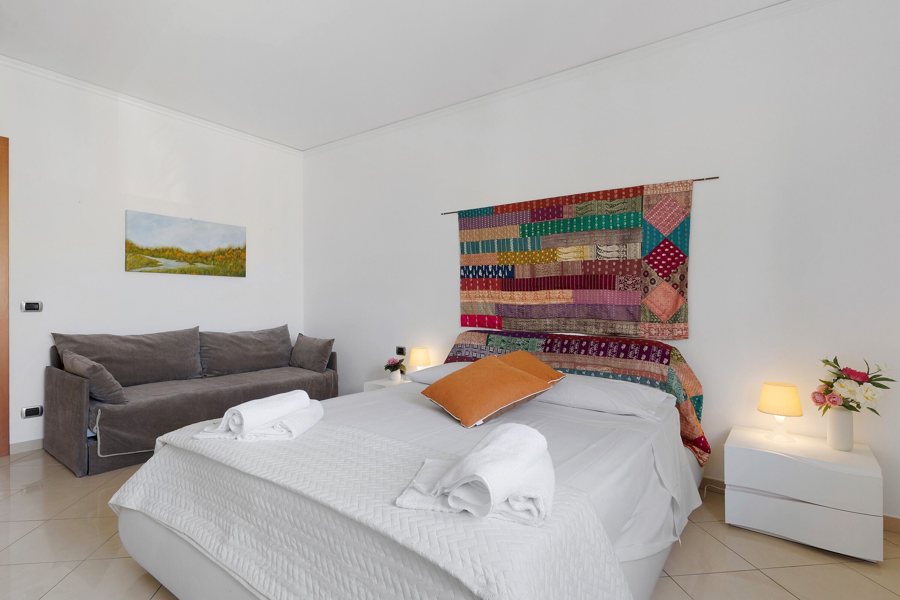 Et hotelværelse med en seng, en sofa, et natbord, en lampe og et vægtæppe.