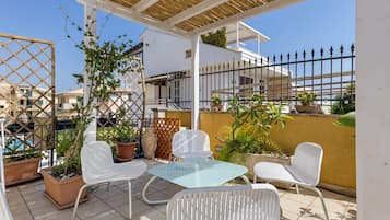 An outdoor patio with white metal chairs and a glass table, surrounded by potted plants and a wooden pergola.