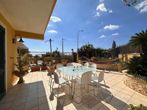 Eine Terrasse mit Glastisch und Stühlen, umgeben von Topfpflanzen und einer Hängematte.