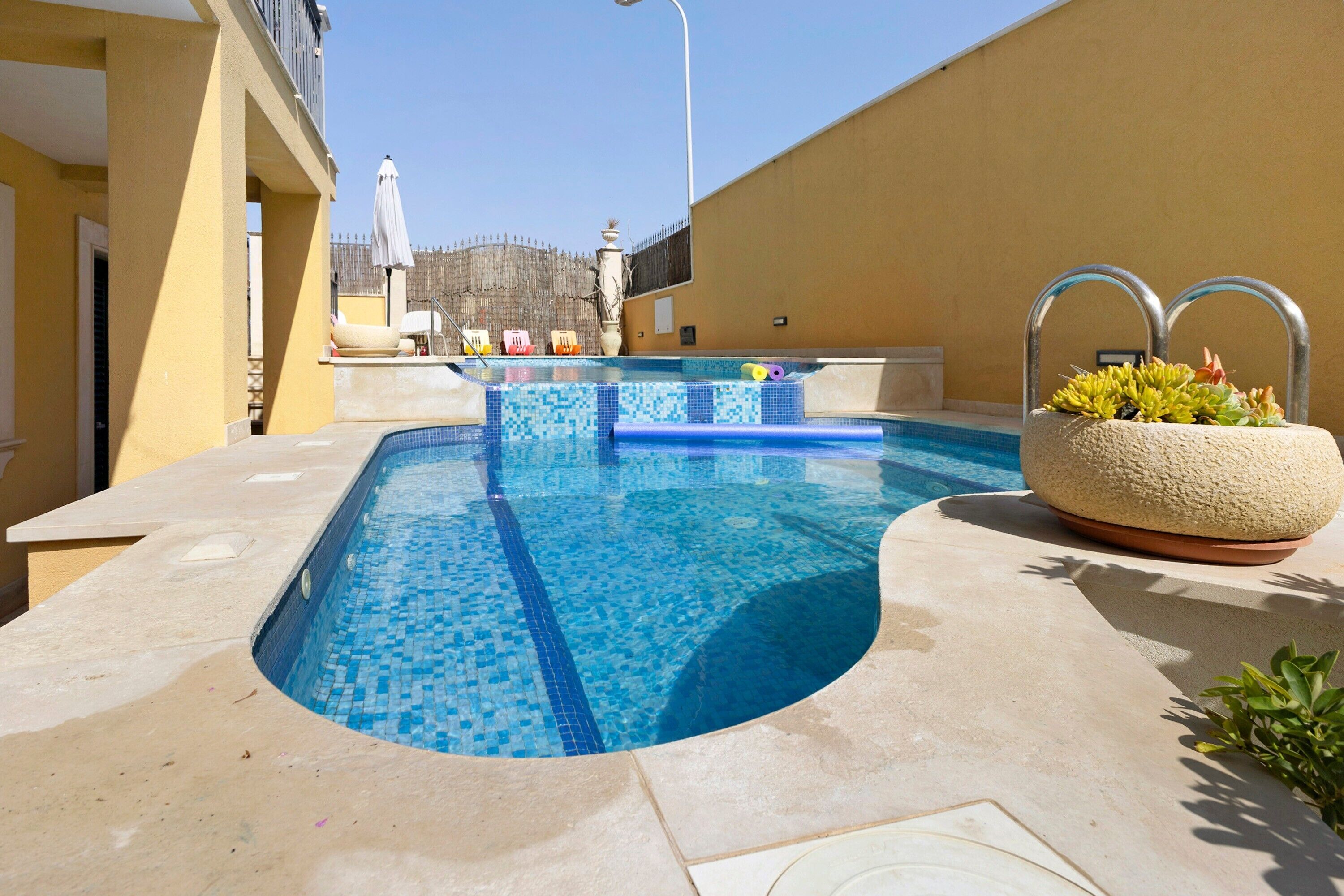 Une piscine bordée de carreaux, un jardinier avec des plantes succulentes et un ciel bleu azur.