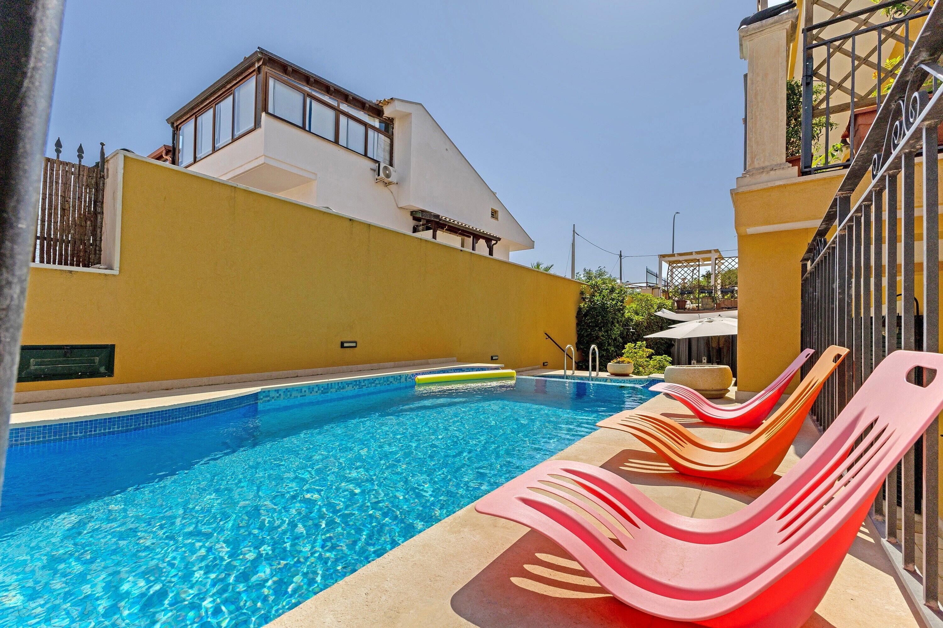 Une piscine à l’eau d’un bleu limpide, entourée d’un mur jaune et d’un bâtiment blanc doté d’un balcon.