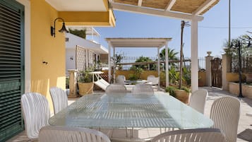 Un espace extérieur abrité, aménagé pour les repas, avec une table en verre et des chaises blanches, entouré de plantes en pot et offrant une vue sur la mer.