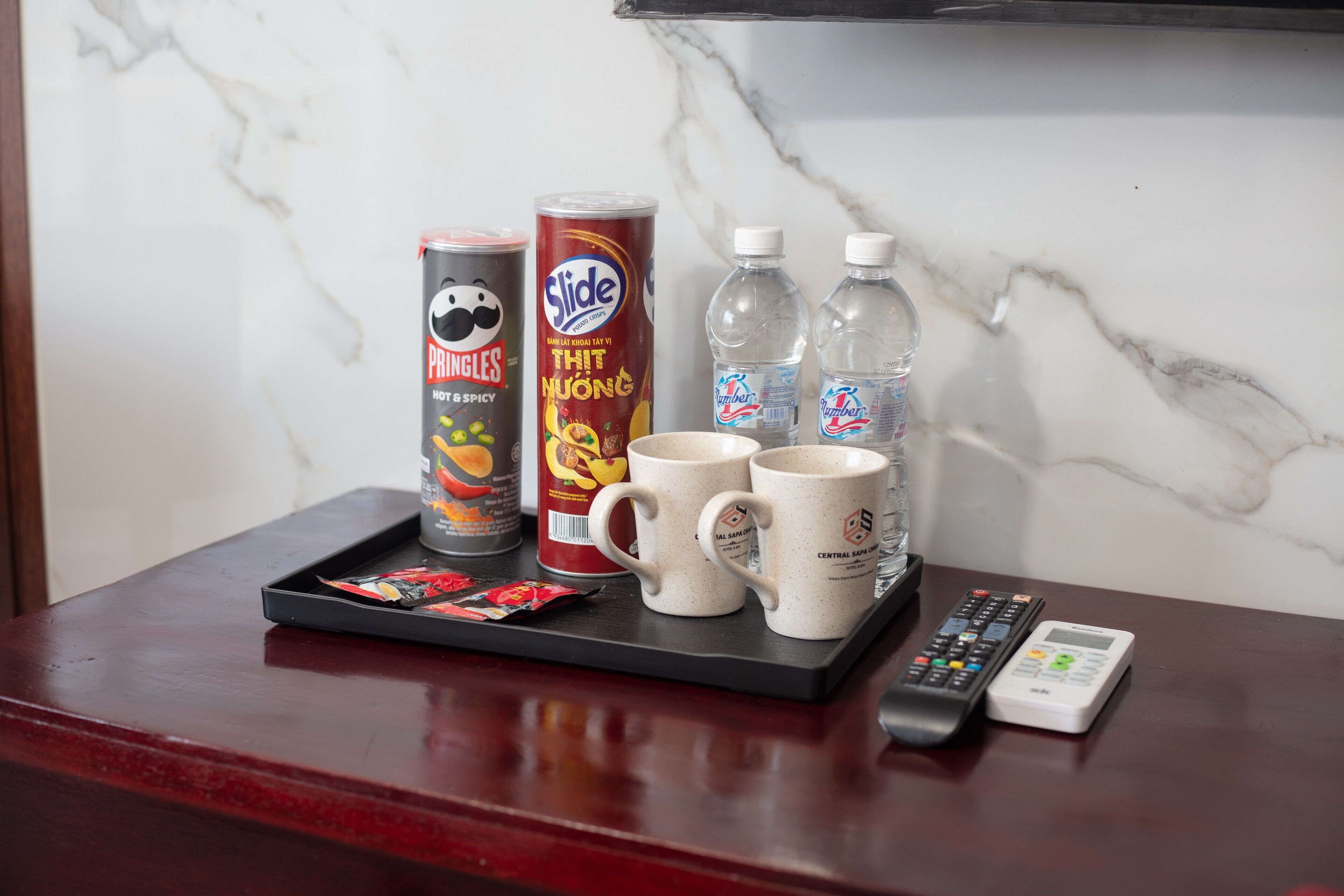 Una bandeja con una lata de Pringles, una lata de Slide, dos botellas de agua, dos tazas de café y un control remoto sobre una mesa de madera.