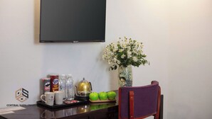 A room with a flat-screen TV mounted on the wall, a table with a tray of green apples, a vase with flowers, and a chair.