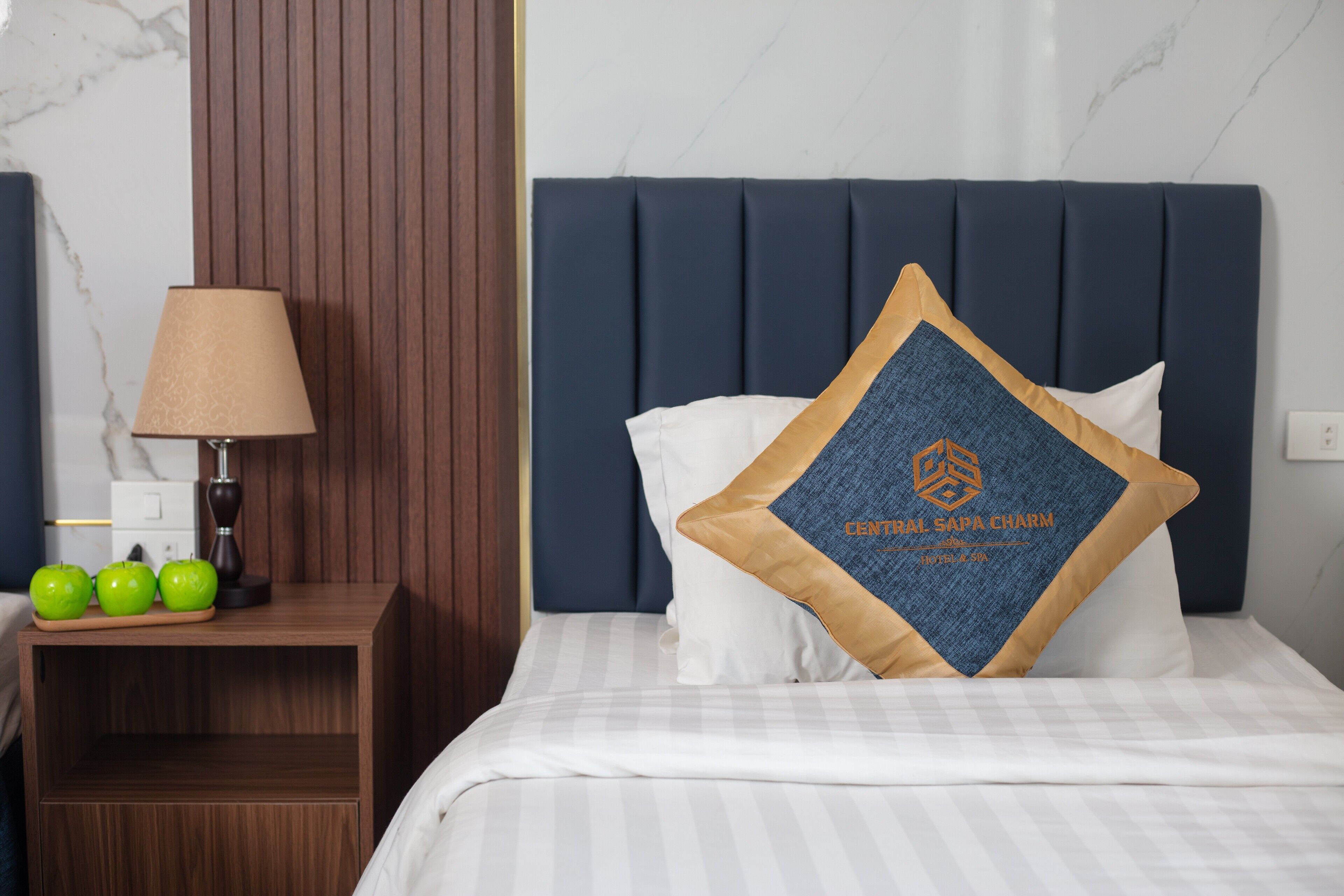A neatly made bed with a blue headboard, a bedside table with a lamp, and three green apples on the table.