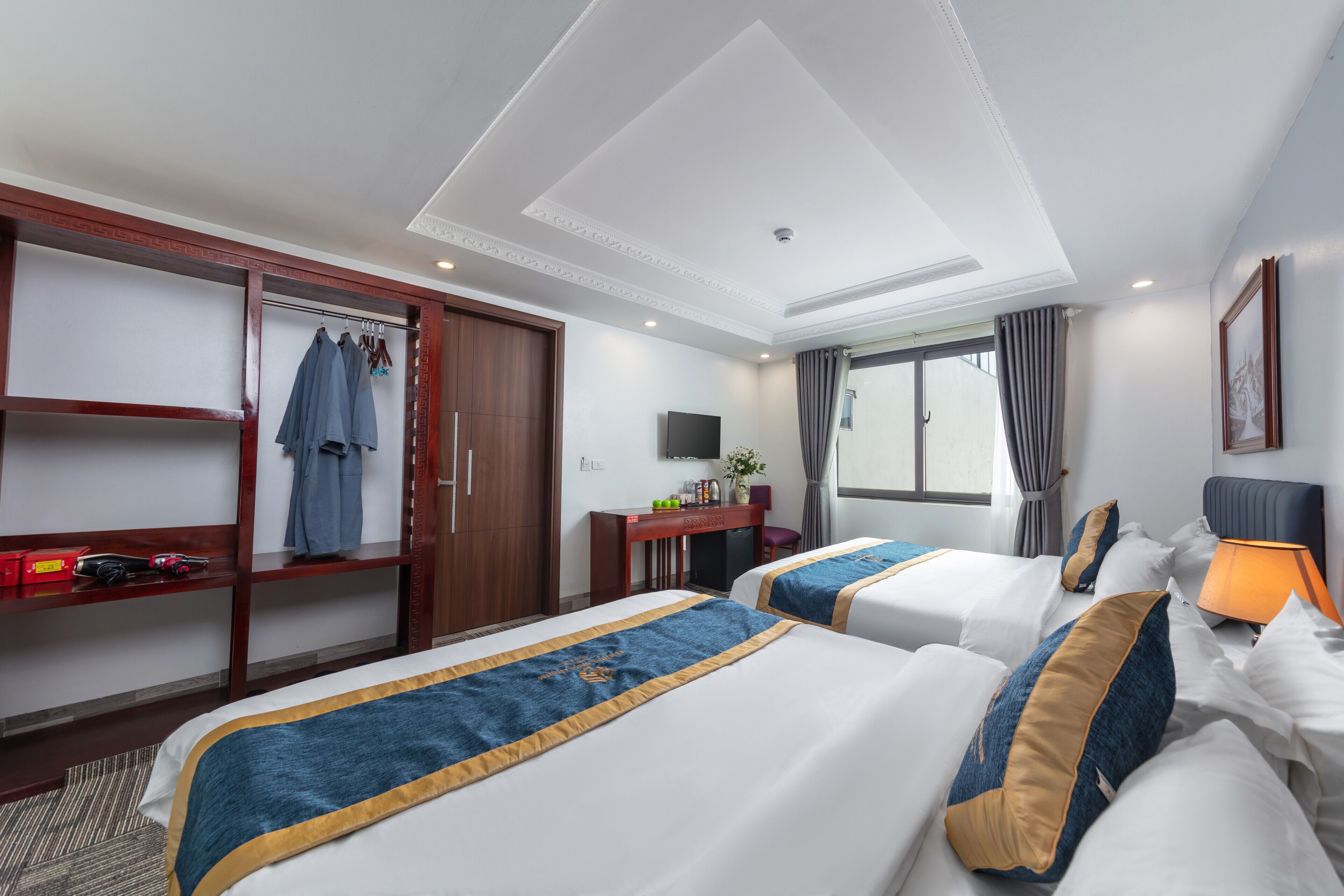 Quarto de hotel com duas camas, uma escrivaninha com televisão, um guarda-roupa e uma janela com cortinas.