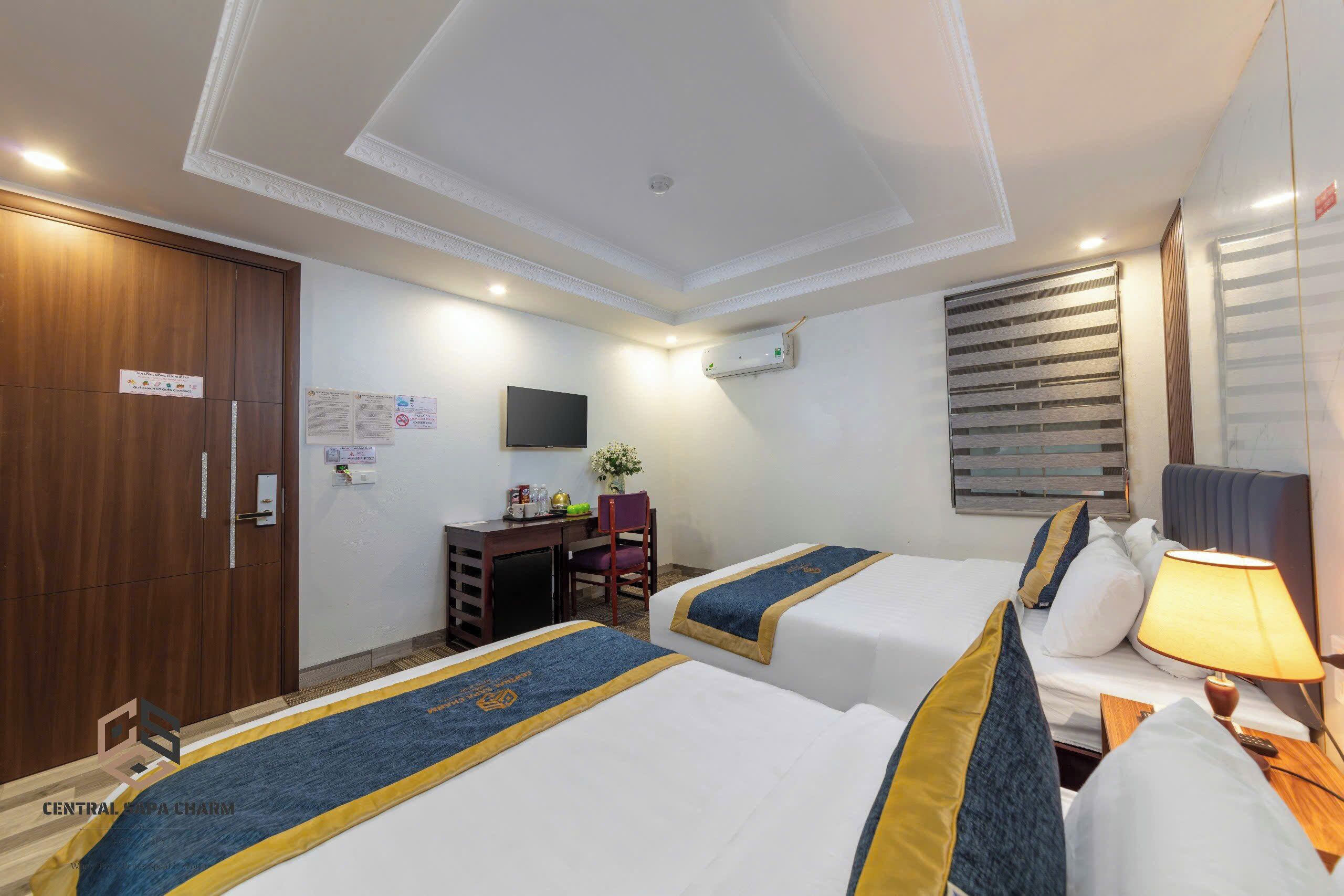 A hotel room with two beds, a desk with a chair, a TV, and an air conditioning unit.