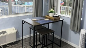 Une pièce avec une table et deux chaises, une plante en pot et une vue sur la rue grâce à de grandes fenêtres.
