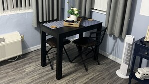 Une pièce comprenant une table, des chaises, une petite plante, un radiateur et une fenêtre avec des rideaux.