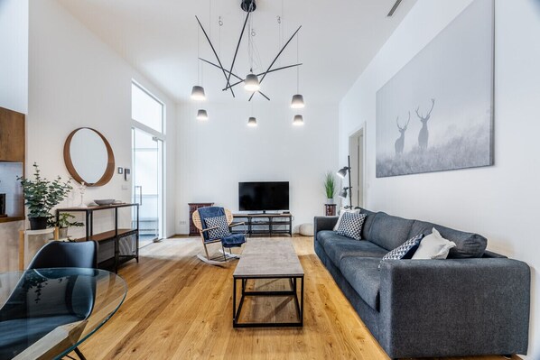 Un salon moderne avec un canapé gris, une table basse en verre et un téléviseur. Le sol est en bois, il y a un miroir rond et une plante.