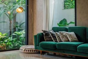 Un canapé vert agrémenté de coussins à motifs, un lampadaire et une vue sur la verdure par une grande fenêtre.