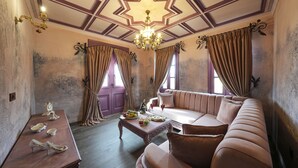 Ein Zimmer mit einer großen Couch, einem Couchtisch mit Früchten und Getränken, einer Kristallleuchtre und einem Holznachttisch mit Schuhen.