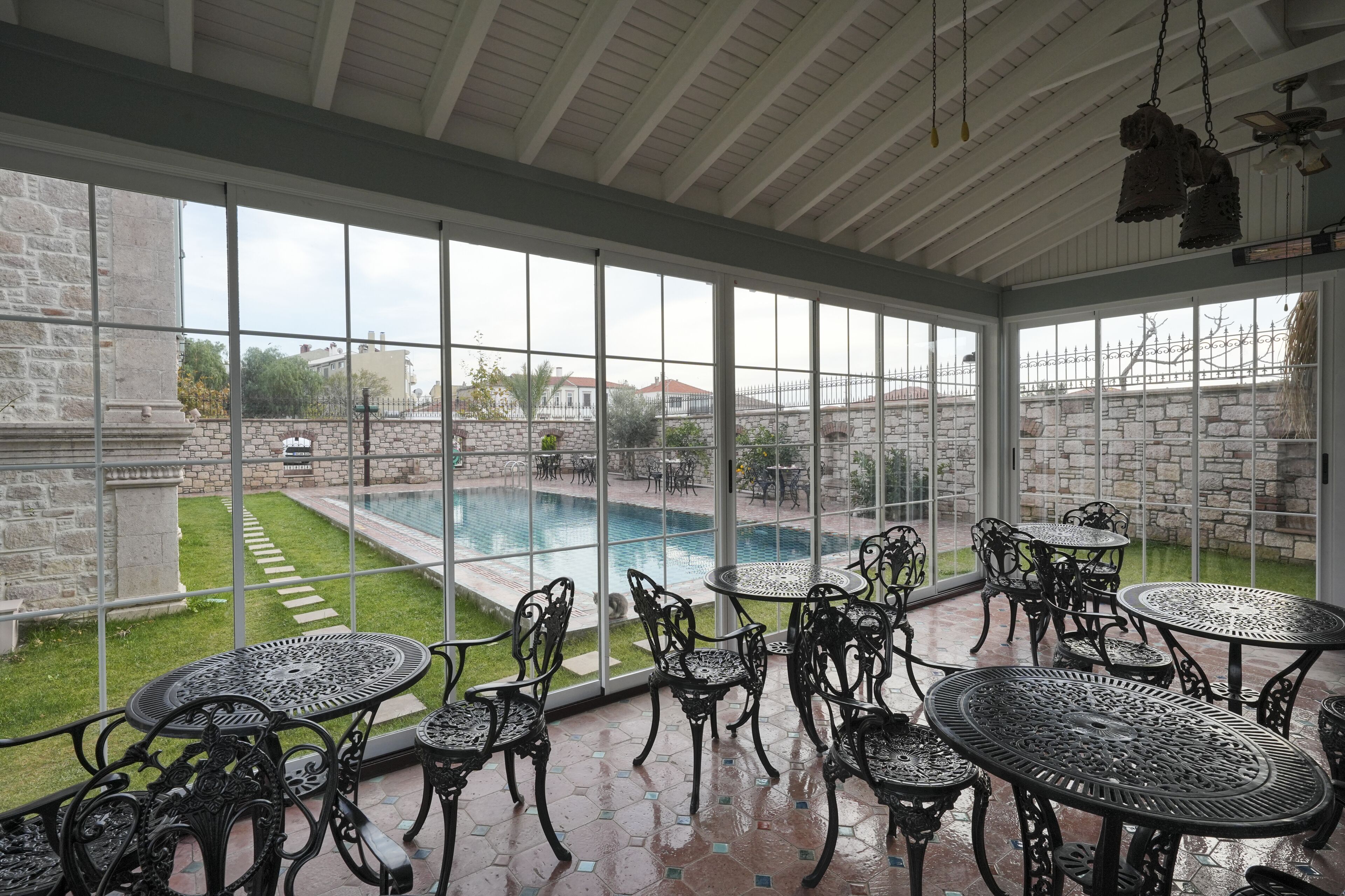 A covered patio with a pool view, featuring ornate metal tables and chairs, stone walls, and a clear glass roof.