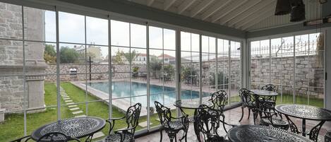 A covered patio with a pool view, featuring ornate metal tables and chairs, stone walls, and a clear glass roof.