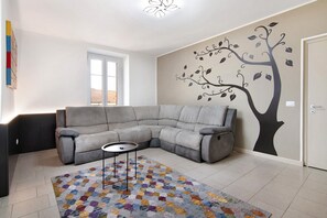 Een woonkamer met een grijze modulaire bank, een kamerkleed met patroon, een muurschildering van een boom en een raam.