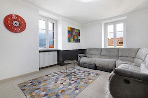 Een woonkamer met een grijze bank, een rode klok, een kleurrijk tapijt en een raam met uitzicht.