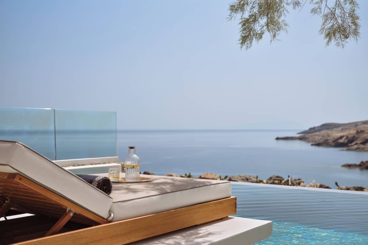 A sun lounger with a bottle and glass on a poolside deck, overlooking a calm sea and rocky coastline.