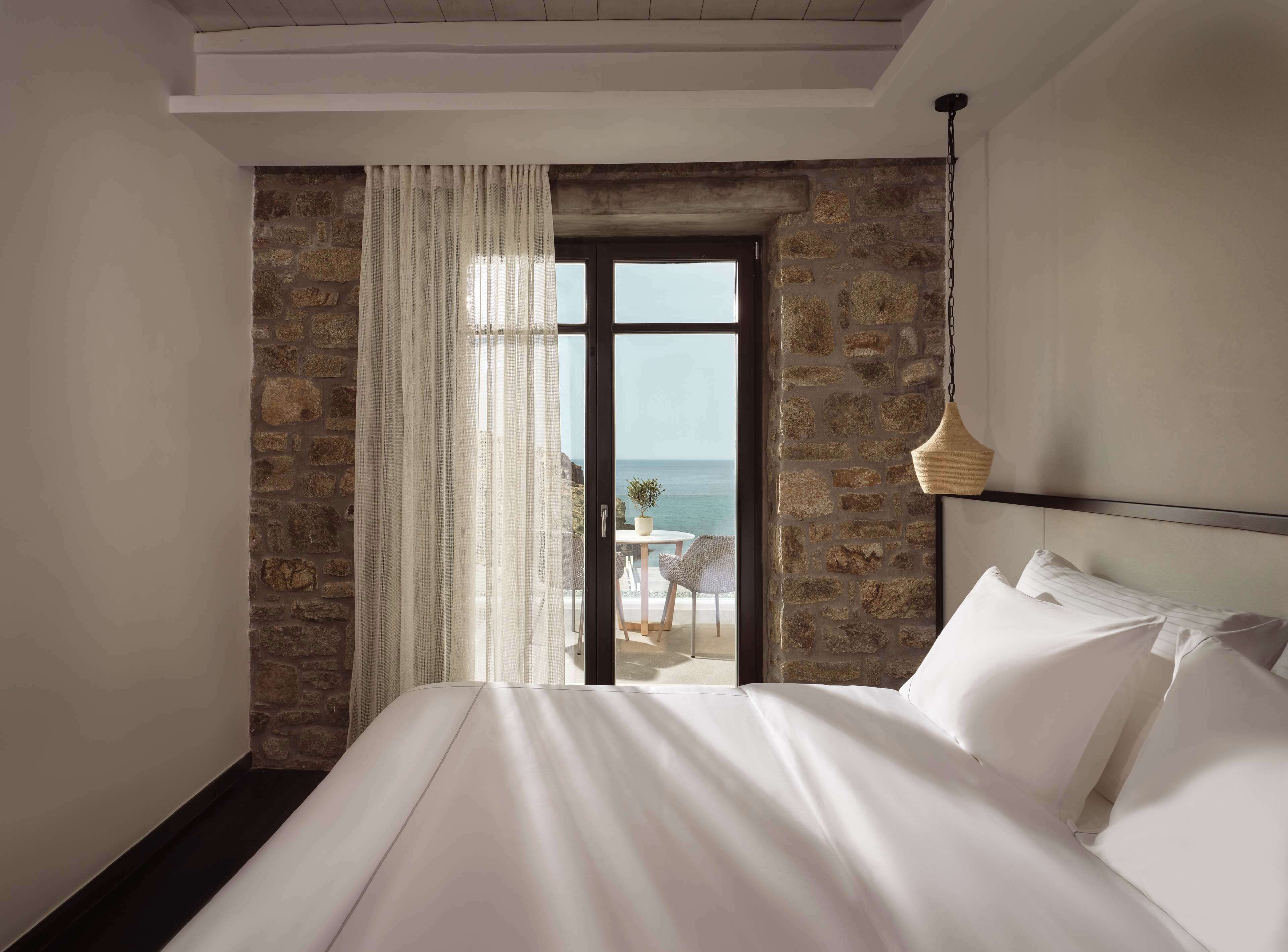A bedroom with a large bed, stone walls, a view of the sea, and a dining area with a table and chairs outside the window.