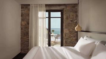 A bedroom with a large bed, stone walls, a view of the sea, and a dining area with a table and chairs outside the window.