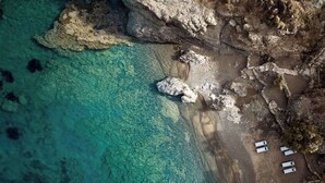 An aerial view of a beach with clear turquoise water, rocky terrain, and several white structures that appear to be part of a resort or hotel. - Semeli Coast Mykonos, Curio Collection by Hilton (Mykonos)