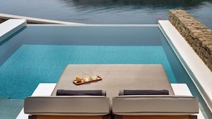 An infinity pool with a view of a rocky coastline. There are lounge chairs and a tray with glasses and a lemon. - Semeli Coast Mykonos, Curio Collection by Hilton (Mykonos)