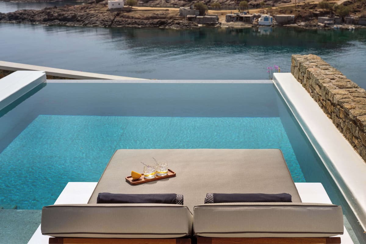 An infinity pool with a view of a rocky coastline. There are lounge chairs and a tray with glasses and a lemon.