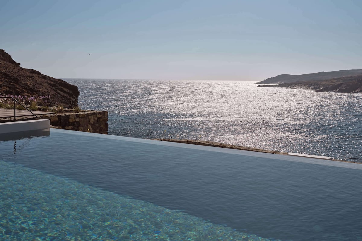 A clear view of a swimming pool with a stone edge, overlooking a bright, calm sea under a clear sky.