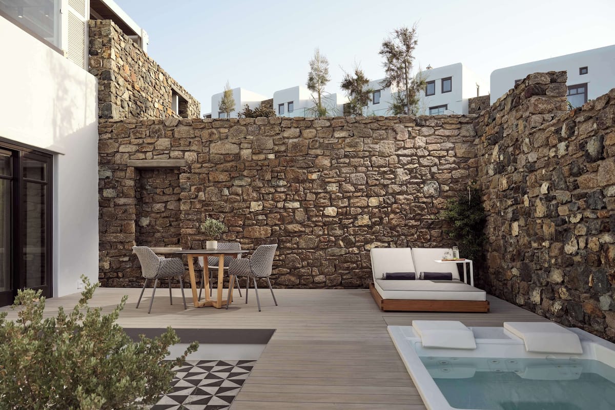 A modern outdoor patio with a stone wall, a wooden deck, a dining area with chairs, a sofa, and a hot tub.