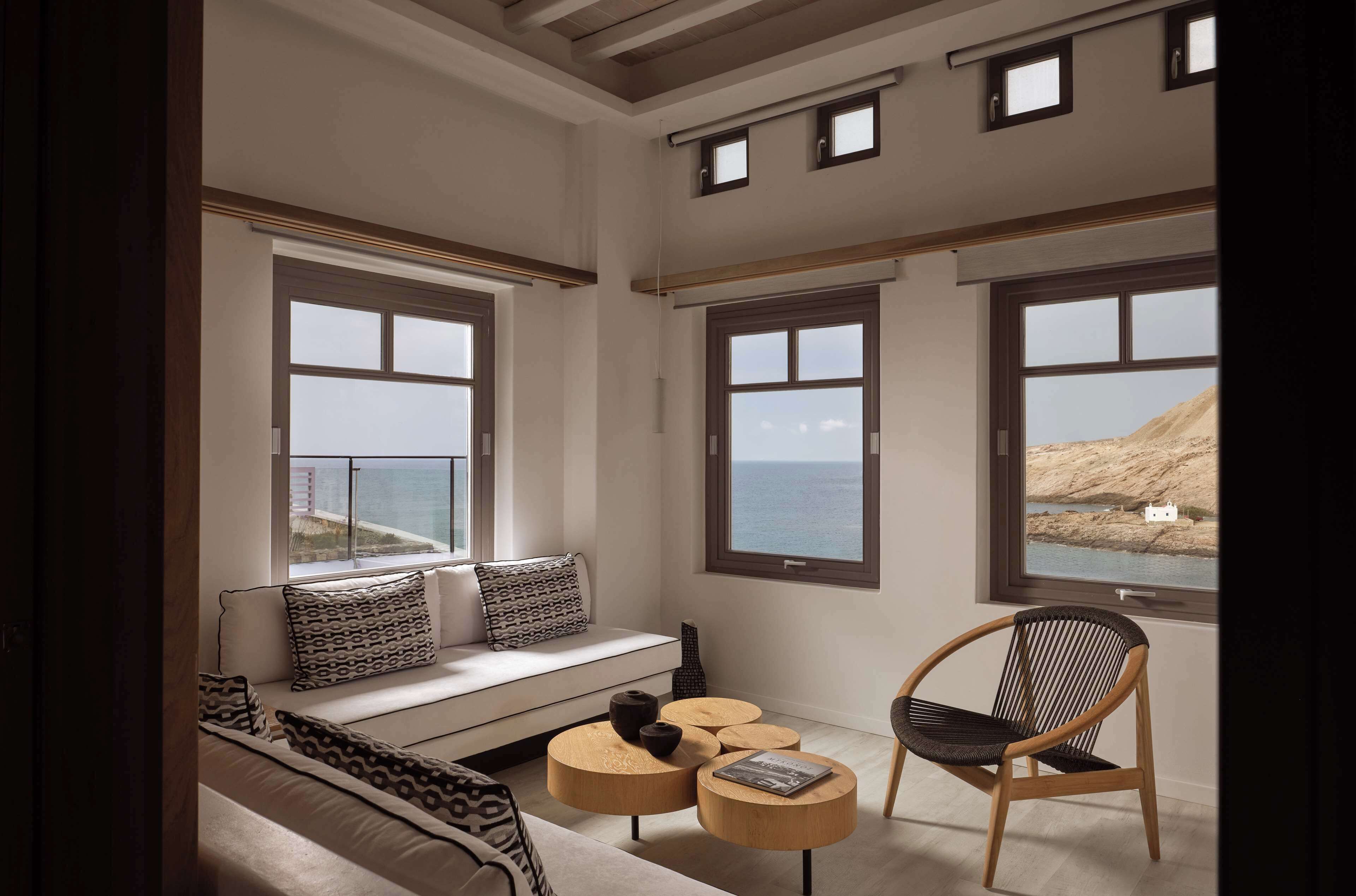 A modern living room with a sofa, a round coffee table, and a chair, large windows with a view of the sea and hills, and a wooden ceiling.